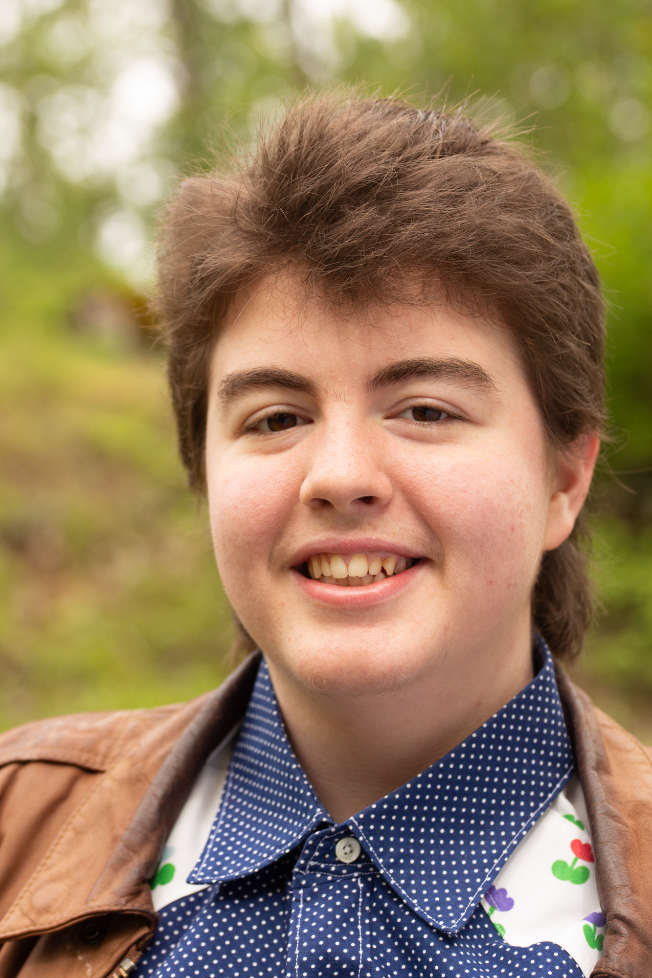 Brooklyn's headshot, wearing a blue collared shirt and a brown leather jacket, smiling.