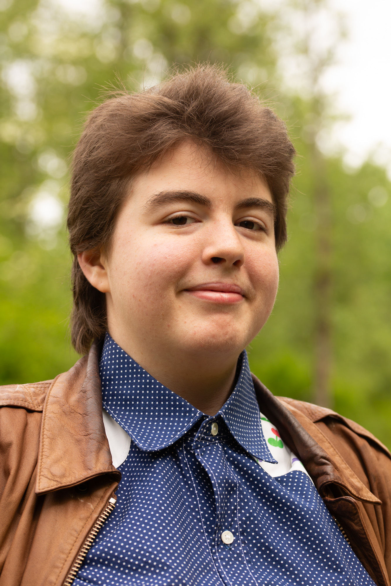 Brooklyn's headshot, wearing a blue collared shirt and a brown leather jacket, smiling.